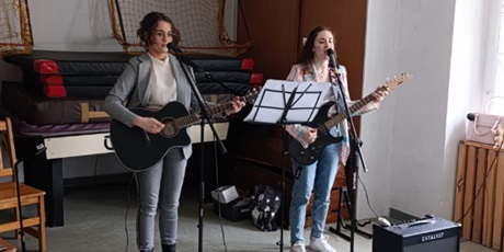 Powiększ grafikę: Dwie wokalistki z gitarami w dłoniach, na małej zaimprowizowanej scenie, w ich otoczeniu znajdują się elementy nagłośnienia.