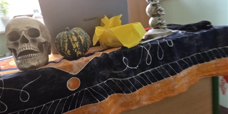 Powiększ grafikę: Na zdjęciu widać biurko lub stolik przykryty ciemno-granatową i pomarańczową tkaniną z ozdobnym wzorem, przypominającym halloweenowy obrus. Na blacie ustawiono kilka dekoracji: po lewej stronie znajduje się model ludzkiej czaszki, obok leży mała, pasiasta dynia w kolorach zielonym i żółtym, a dalej widoczny jest żółty papierowy element – prawdopodobnie część ozdoby lub rekwizyt z zabawy. Po prawej stronie stoi świecznik złożony z kilku ułożonych na sobie czaszek.
