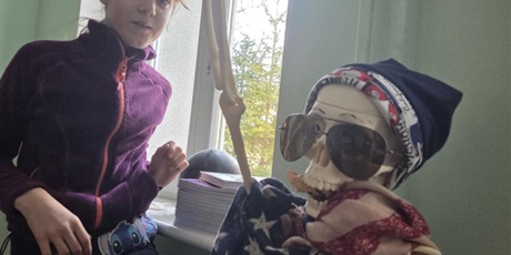 Powiększ grafikę: Na zdjęciu widać model szkieletu ludzkiego przystrojony w zabawny, halloweenowy sposób. Szkielet ma na sobie czapkę, okulary przeciwsłoneczne i jest owinięty apaszką w gwiazdy i paski, co nadaje mu humorystyczny wygląd. Obok niego stoi uczennica, która z zainteresowaniem przygląda się scenie. W kadrze widać też czyjąś rękę trzymającą plastikową dłoń szkieletu, jakby ktoś próbował „podawać mu rękę”.