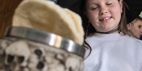 Powiększ grafikę: Na zdjęciu widać dziewczynkę ubraną w halloweenowy strój czarownicy – ma na głowie czarny, spiczasty kapelusz ozdobiony wzorkami i uśmiecha się z zamkniętymi oczami. W tle widać fragment klasy, z zieloną tablicą dekoracyjną i drewnianymi drzwiami. Na pierwszym planie znajduje się kubek lub puchar ozdobiony motywem czaszek, w którym leży coś jasnego – prawdopodobnie rekwizyt lub element zabawy.