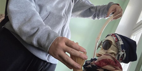 Powiększ grafikę: Na zdjęciu widać ucznia w szarej bluzie z plecakiem, który trzyma za rękę szkielet przystrojony w halloweenowym stylu. Szkielet ma na sobie czapkę, okulary przeciwsłoneczne i apaszki w gwiazdy i paski, co nadaje mu zabawny, niemal modowy wygląd. Uczeń wygląda, jakby prowadził z nim żartobliwą interakcję.