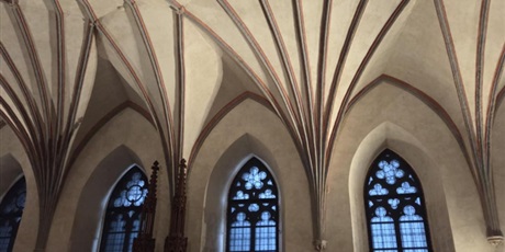 Powiększ grafikę: Fotografia wnętrza kościoła w stylu gotyckim z wysokim, żebrowanym sklepieniem i beżowymi i czerwonymi liniami. Trzy wysokie, łukowate witraże z czarnym maswerkiem w ramę drewniane organy po lewej stronie. Przyciemnione, ambientowe oświetlenie podkreśla detale architektoniczne.