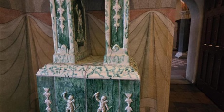 Powiększ grafikę: Fotografia ozdobnej, zielono-białej marmurowej chrzcielnicy z misternymi rzeźbami postaci, umieszczonej w historycznym, słabo oświetlonym kościele. Tło zdobią łukowate drzwi, okładaną tkaninę i kafelkową podłogę, co dodaje architektonicznego bogactwa.